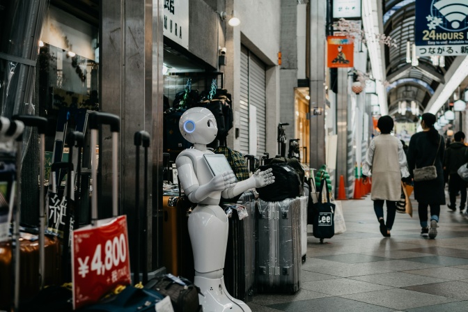 Ein weißer Roboter mit angewinkelten Armen steht am Rand einer Einkaufsstraße. Die Straßenschilder sind mit asiatischen Schriftzeichen beschriftet.