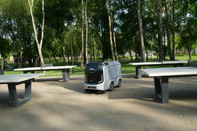 Ein kleines, selbstfahrendes Fahrzeug fährt durch einen Park, vorbei an mehreren Tischtennisplatten aus Beton.