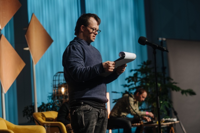Ein Mann mit Down-Syndrom steht auf einer Bühne und liest einen Text von einer Klemmmape ab.