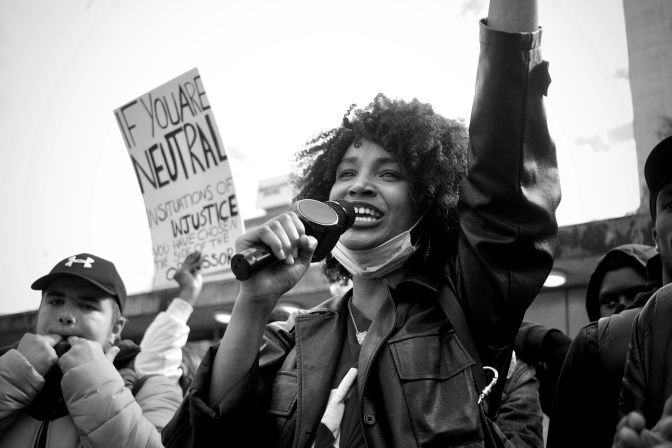 Schwarz-weiß-Foto einer schwarzen Frau, die bei einer Demo in ein Mikro spricht. Sie reckt ihre Faust in die Luft.