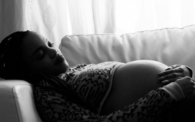 Schwarz-weiß Foto einer schwangeren schwarzen Frau.
Sie liegt auf dem Sofa, ihre Hände auf dem Bauch verschränkt.