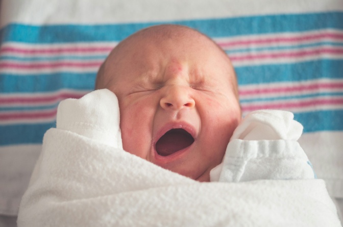 Ein Baby liegt auf einer gestreiften Decke und gähnt mit geschlossenen Augen und weit geöffnetem Mund.