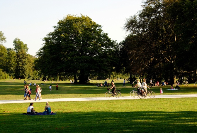 Menschen sitzen in einem Park auf der Wiese, laufen durch oder fahren Fahrrad.