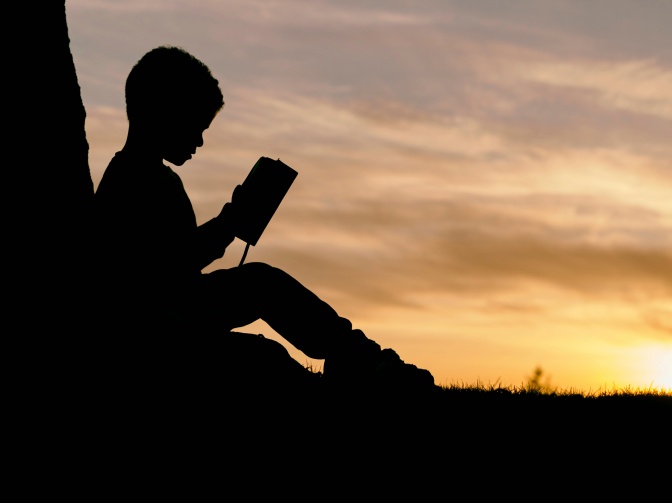 Ein Junge sitzt bei Sonnenuntergang im Freien und liest ein Buch. Er ist nur als Umriss im Gegenlicht zu sehen.