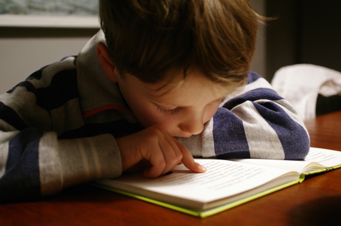 Ein Junge im Grundschulalter sitzt am Tisch und liest ein Buch. Mt dem Finger fährt er beim Lesen die Zeilen entlang.