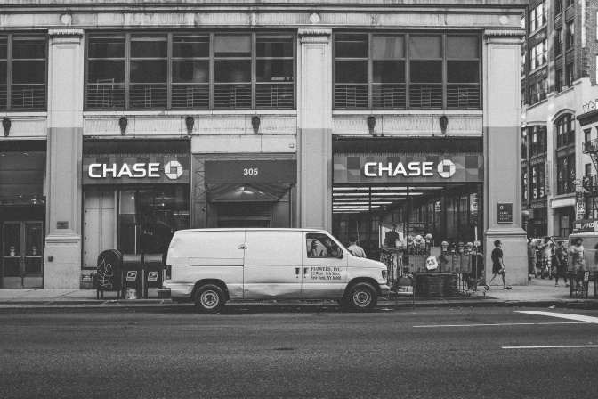 Schwarz-weiß-Foto eines verbeulten, weißen Transporters, der vor einer Bank parkt.