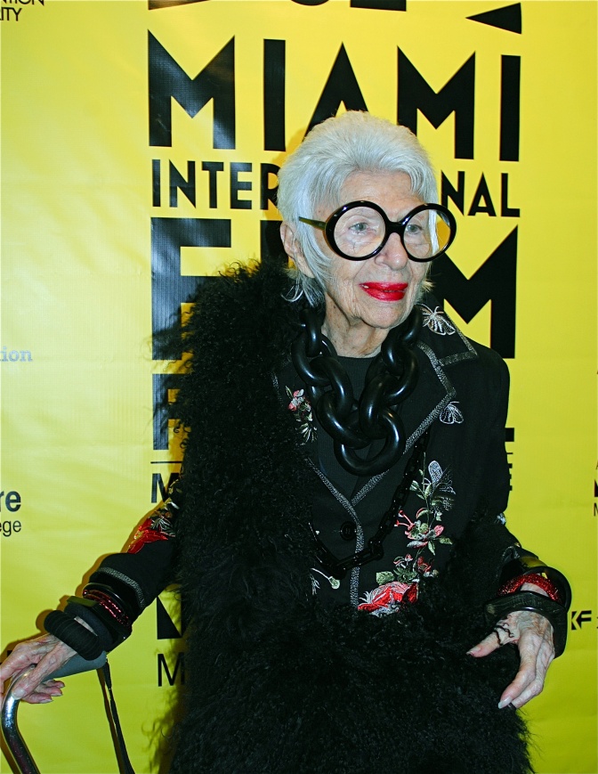 Iris Apfel mit kurzen, weißen Haaren mit großer schwarzer Brille und in schwarzer Kleidung. Sie steht vor einer Logowand eines Filmfestivals in Miami.
