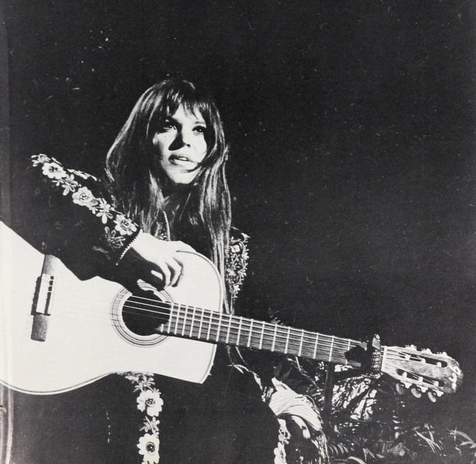 Schwarz-weiß-Foto einer jungen Frau mit Gitarre und Lederjacke auf der Bühne.