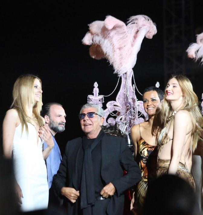 Roberto Cavalli auf dem Laufsteg zwischen mehreren Models. Im Hintergrund ein riesiger, glitzernder Kronleuchter.