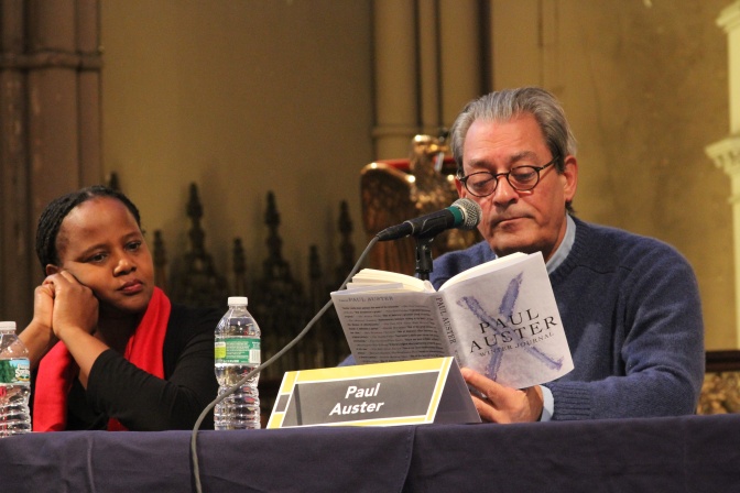 Paul Auster liest auf einer Bühne aus einem seiner Bücher vor. Neben ihm sitzt eine schwarze Frau und hört konzentriert zu.