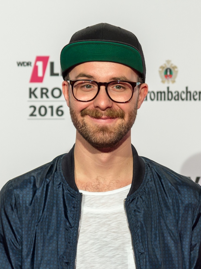 Mark Forster mit Vollbart, Brille und Schirmmütze vor einer Logowand der Einslive Krone