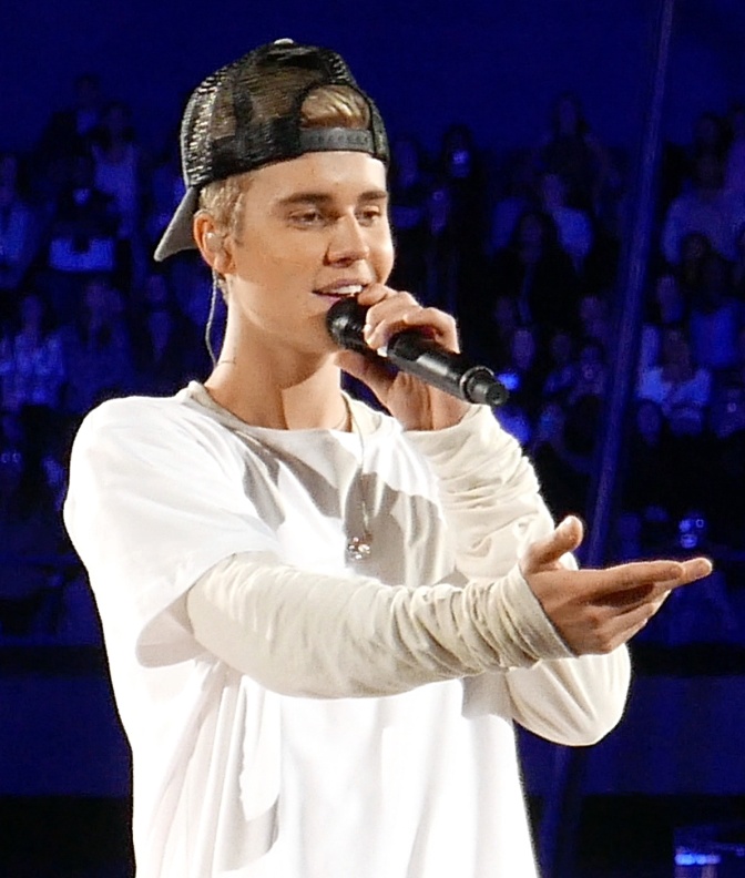 Justin Bieber singt auf der Bühne i ein Mikro ud streckt den linken Arm aus. Er trägt weiße Kleidung und eine schwarze, nach hinten gedrehte Schirmmütze.