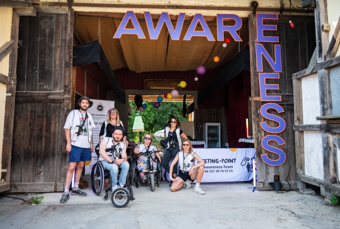 6 Personen in weißen Warnwesten, zwei davon im Rollstuhl. Sie stehen vor einem Gebäude mit dem Schriftzug Awareness.