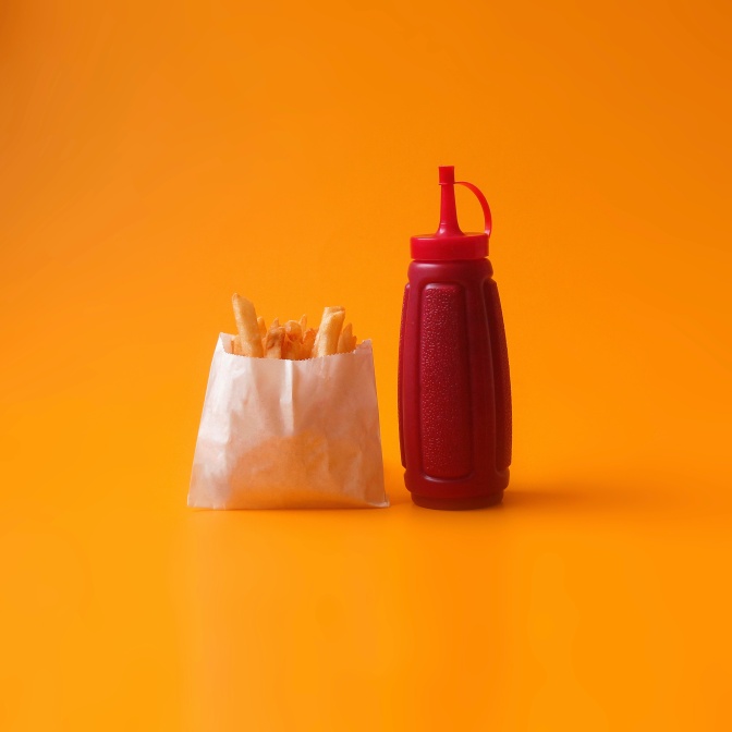 Eine kleine Portion Pommes in einer weißen Papiertüte, daneben eine Flasche Ketchup.