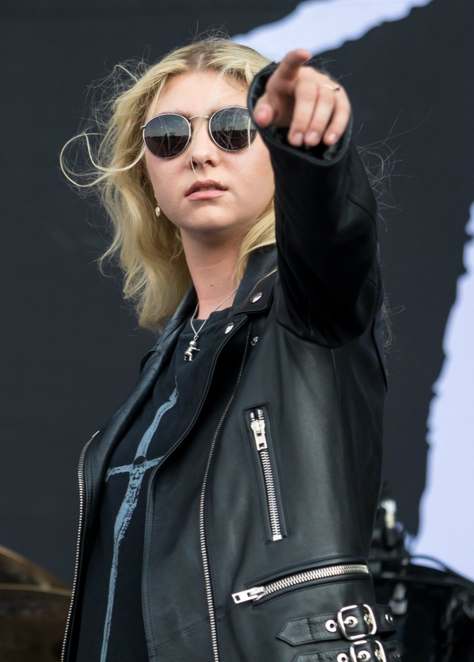 Taylor Momsen in schwarzer Lederkleidung und mit schwarzer Sonnenbrille. Sie zeigt mit ausgestrecktem Arm in Richtung der Kamera.