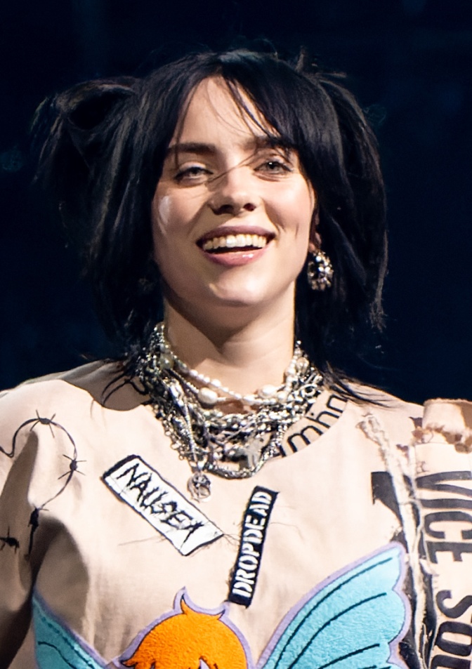Billie Eilish mit schwarzen Haaren in einem gemusterten Shirt auf der Bühne. Sie lächelt offen in die Kamera.