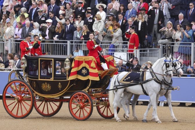 Eine Kutsche wird von 2 weißen Pferden gezogen. In der Kutsche sitzt Prinzessin Kate. Im Hintergrund sitzen Zuschauer*innen in Rängen.