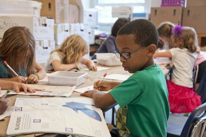 Schüler*innen malen in einem Klassenraum Bilder vo Flugzeugen aus. Im Vordergrund ist ein schwarzer Junge mit Brille im Bild zu sehen.