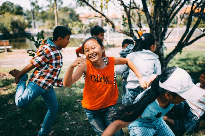 Ein asiatisches Mädchen rennt und lacht. Um sie herum ist eine Gruppe anderer spielender und rennender Kinder.
