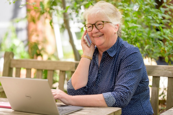 Eine ältere Frau telefoniert man Handy und lächelt breit. Vor ihr steht ein aufgeklappter Laptop.