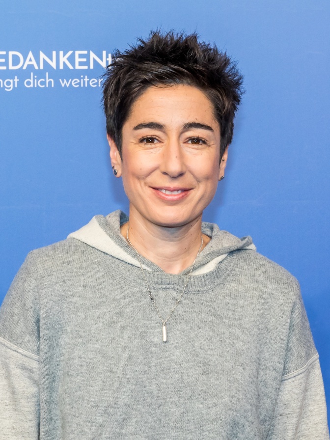 Dunja Hayali in einem rauen Pullover mit kurzen, nach oben frisierten Haaren vor einer blauen Logowand.