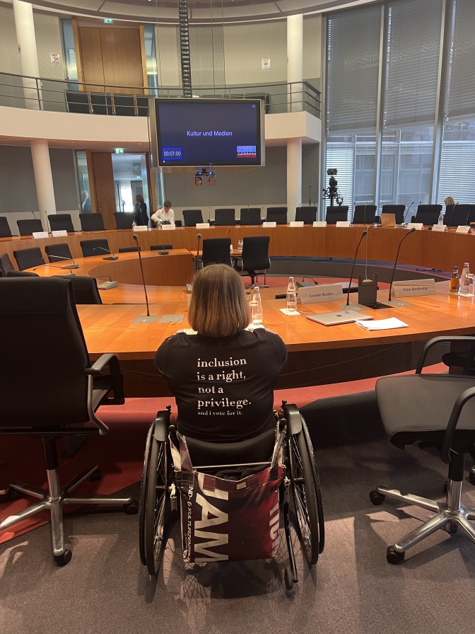 Franziska Lammers an einem großen runden Tisch mit Namensschildern. Die Stühle sind noch unbesetzt. Sie trägt ein schwarzes T-Shirt mit dem Aufdruck: INclusion is a right, not a Privilege. And I vote for it.