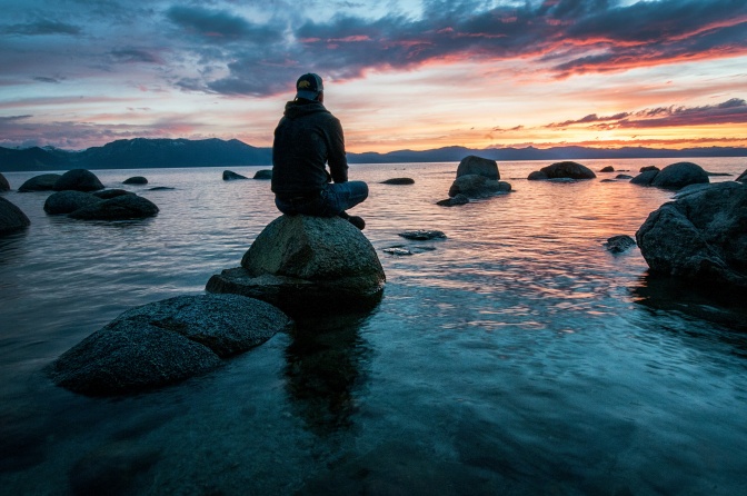 Ein Mann mit Schirmmütze sitzt im Wasser auf einem großen Stein und meditiert bei Sonnenuntergang.