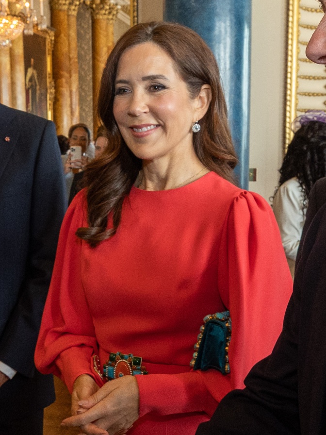 Mary von Dänemark mit braunen, langen, gewellten Haaren in einem festlichen roten Kleid. Sie lächelt.