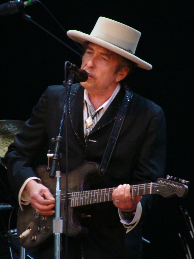 Bob Dylan steht vor einem Mikro, spielt Gitarre und singt. Er trägt einen Anzug und einen weißen Hut.