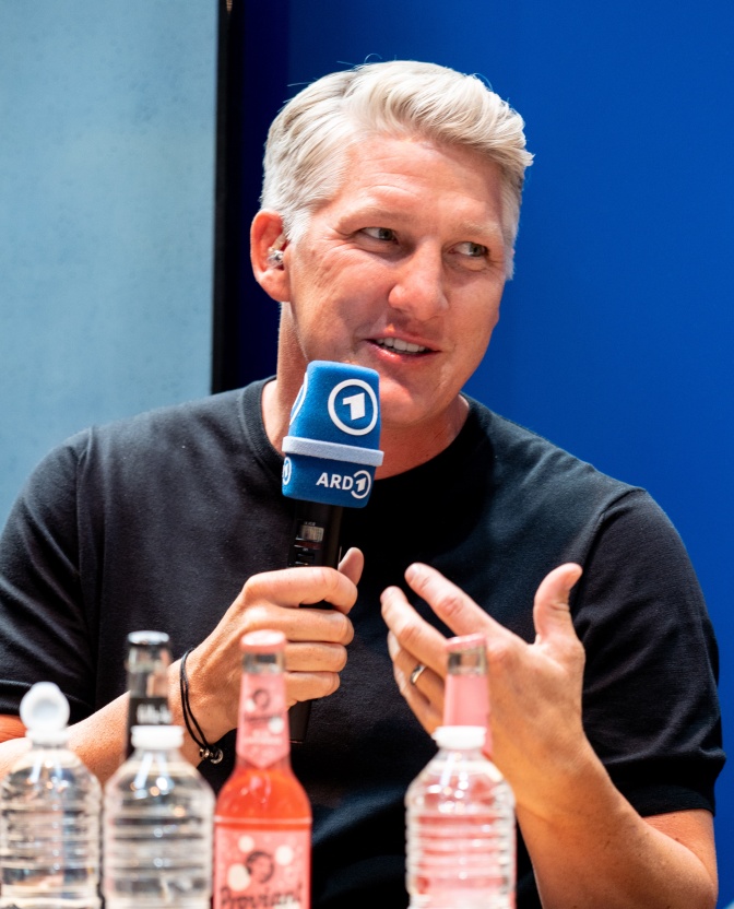 Bastian Schweinsteiger sitzt an einem Tisch, spricht in ein Mikro und gestikuliert mit der Hand.