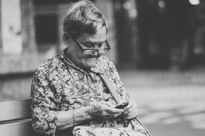 Schwarz-weiß-Foto einer alten Frau mit kurzen Haaren und Brille. Sie sitzt auf einer Bank und benutzt ein Handy.