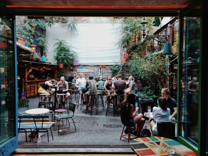 Menschen sitzen in einem Innenhof in einem Biergarten. Das Foto ist vom Innenraum aus durch eine geöffnete Glastür fotografiert.