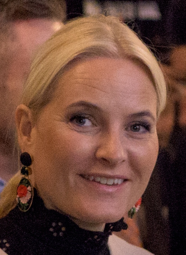 Mette-Marit mit blonden Haaren in einer schwarzen Spitzenbluse mit großen Ohrringen.