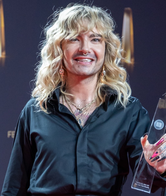 Bill Kaulitz mit gelockten. blondierten Haaren. Er trägt ein schwarzes Hemd und lächelt in die Kamera. Erhält den Deutschen Fernsehpreis in der Hand.