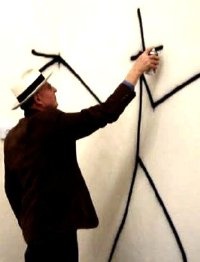 Harald Naegeli in schwarzem Anzug mit weißem Hut. Er sprayt eine Figur aus schwarzen Linien an eine weiße Wand.