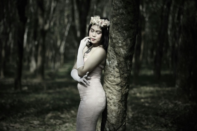 Schwarz-weiß-Foto einer Frau im eleganten Kleid und mit Blumenkranz, die mit geschlossenen Augen an einem Baum lehnt.