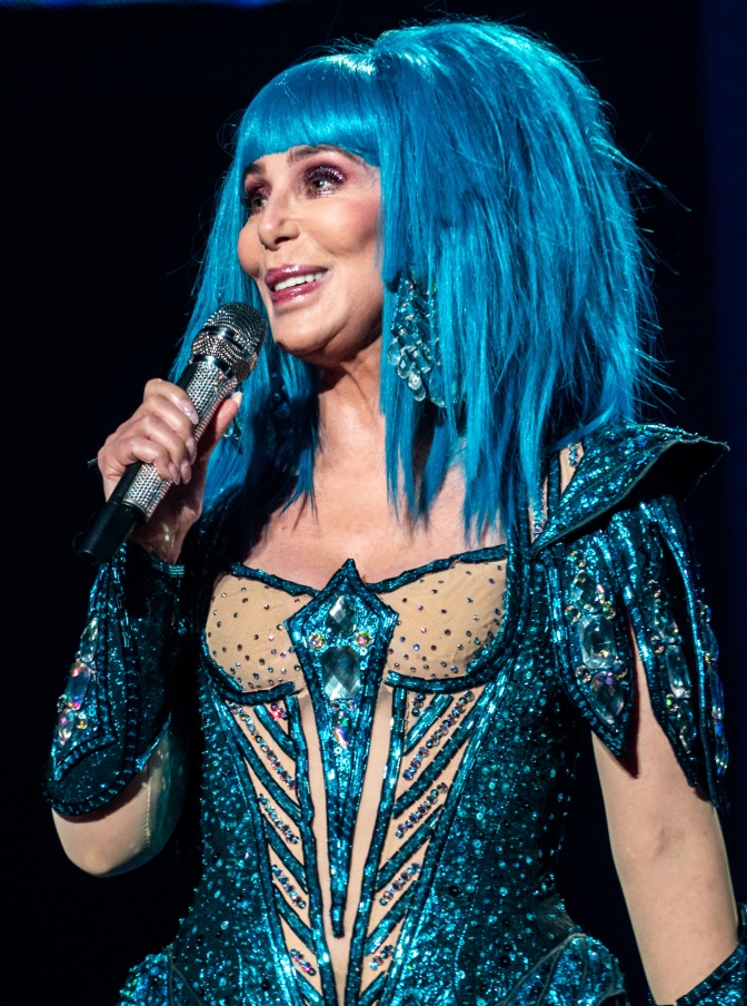 Cher in einem aufwendigen Bühnenkostüm mit großer blauer Perücke. Sie singt in ein Mikrofon.