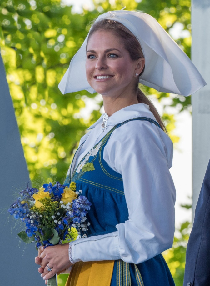 Madeleine von Schweden in schwedischer Tracht mit einer großen weißen Haube und einem Strauß Blumen in der Hand. Sie lächelt.