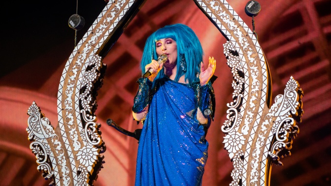 Cher in einem Bühnenkostüm in blau mit blauer Perücke. Sie singt in ein Mikro.