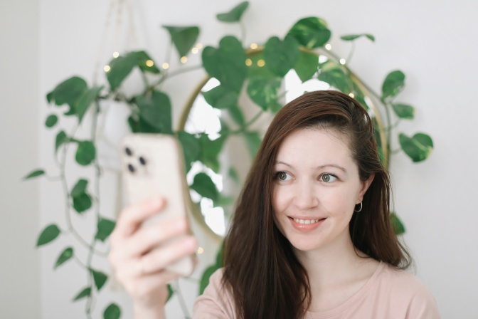 Eine Frau, die nackt zu sein scheint, fotografiert sich selbst mit dem Handy. Hinter ihr sieht man eine große Zimmerpflanze.