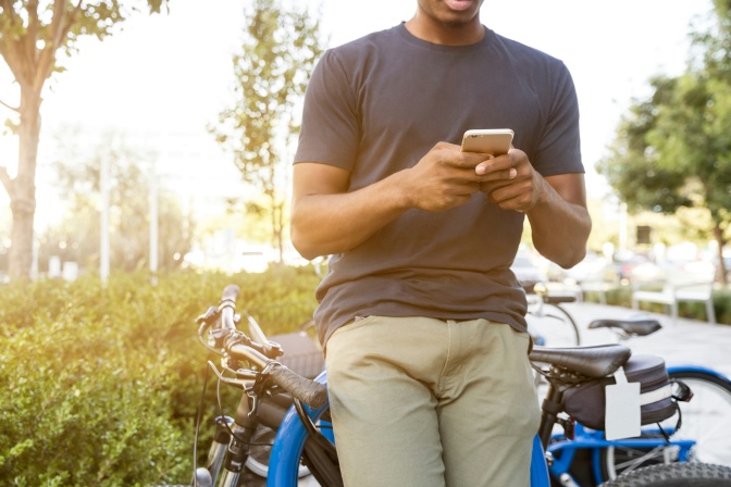 Ei Mann lehnt an einem Fahrrad und hat ei Handy in beiden Händen.