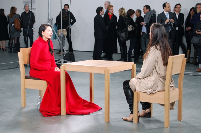 Zwei Frauen sitzen sich an einem Tisch gegenüber und sehen sich an. Im Hintergrund steht Publikum und betrachtet die Performance.