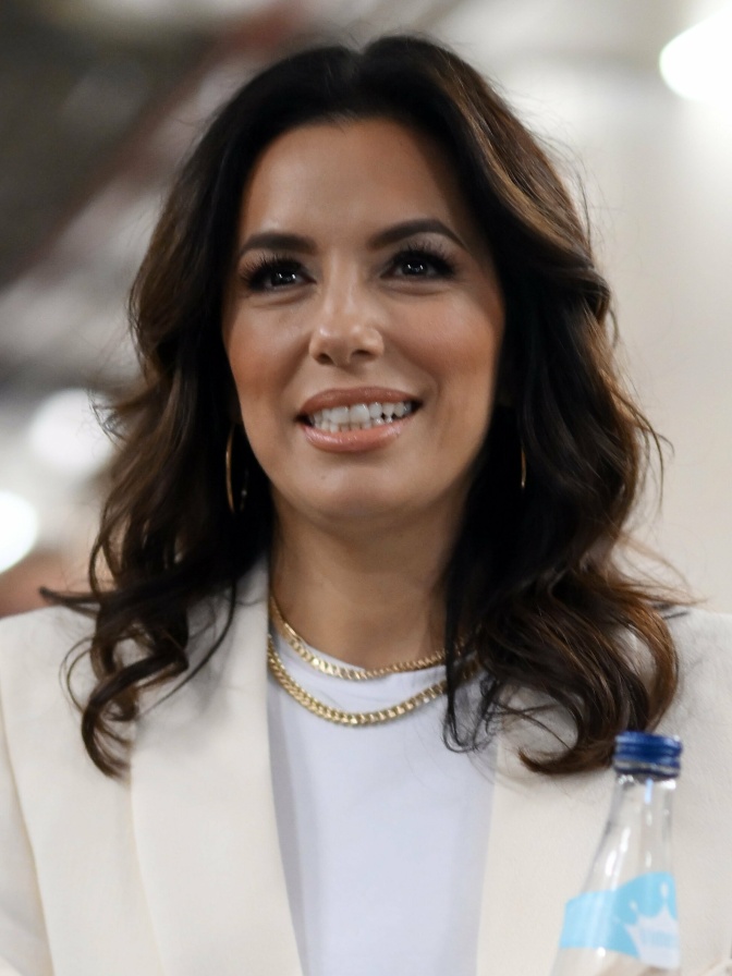 Eva Longoria mit gewellten braunen Haaren. Sie trägt ein weißes Sakko und schaut lächelnd in die Kamera.