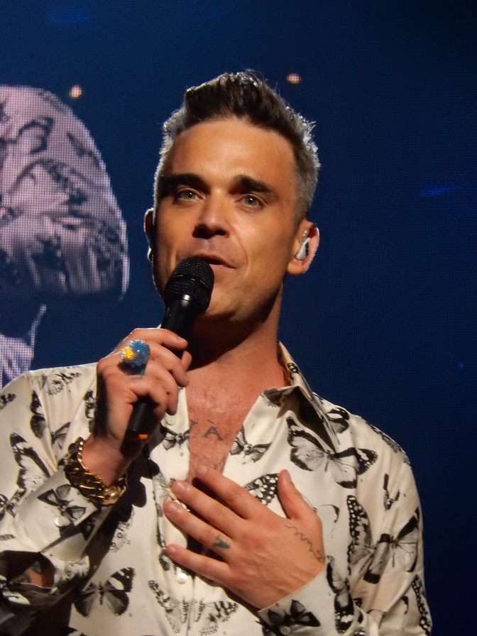 Robbie Williams hält ein Mikro in der Hand und singt. Er trägt ein Hemd mit Schmetterlingsmuster.