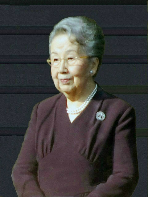 Eine alte japanische Dame in einem dunkelroten Kleid. Die grauen Haare hat sie hochgesteckt. Sie trägt Brille und Perlenkette.