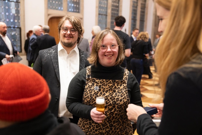 Natalie Dedreux mit einem Glas Kölsch in der Hand, einer ihr steht ein Mann mit Down-Syndrom im Anzug.