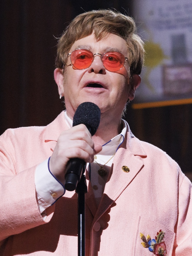 Elton John in einem hellrosanen Sakko mit runder, orangefarbener Brille. Er singt in ein Mikrofon.