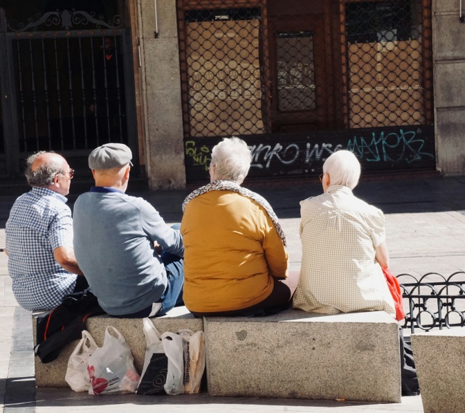 Vier ältere Menschen sitzen auf Steinbänken und unterhalten sich. Sie sind auf dem Foto von hinten zu sehen.