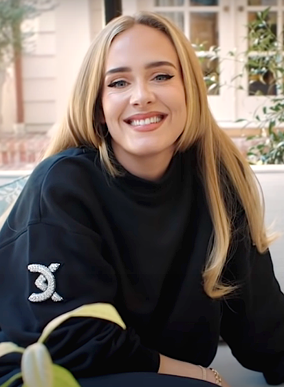 Adele in einem schwarzen Sweatshirt mit Chanel-Logo am Ärmel. Sie hat lange, glatte blonde Haare und lächelt direkt in die Kamera.