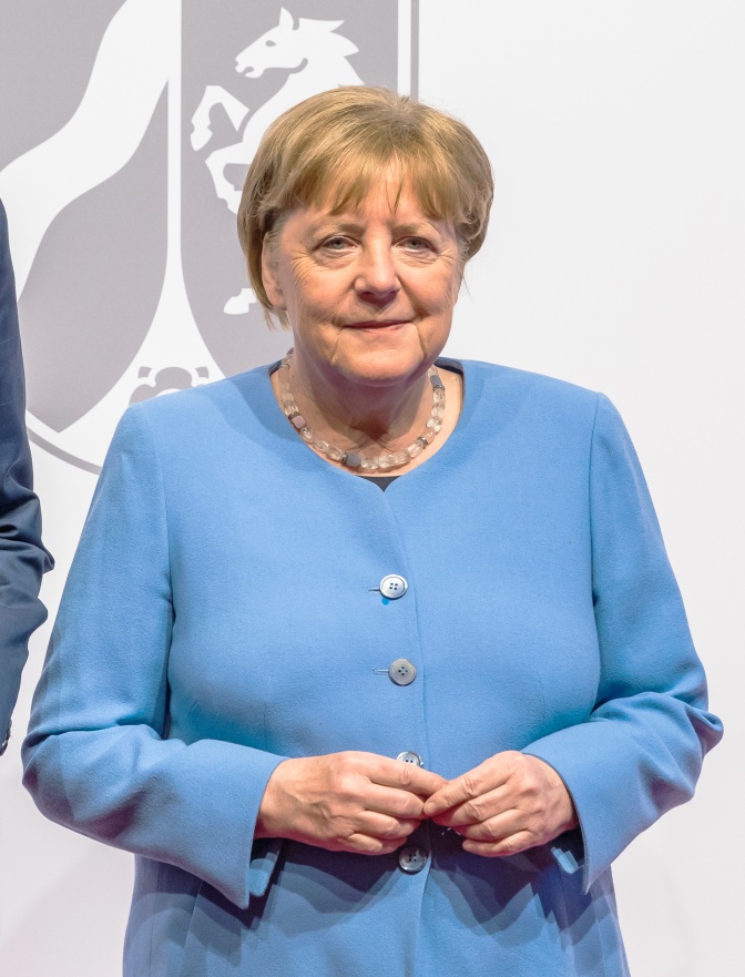 Angela Merkel in einem hellblauen Kostüm. Sie steht vor einem grauen Wappen und hat die Hände vor dem Körper zusammengelegt.